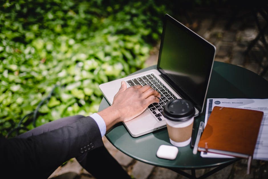 High angle of crop anonymous young man in formal suit typing on laptop while sitting at round table with coffee notebook and documents in outdoor cafe