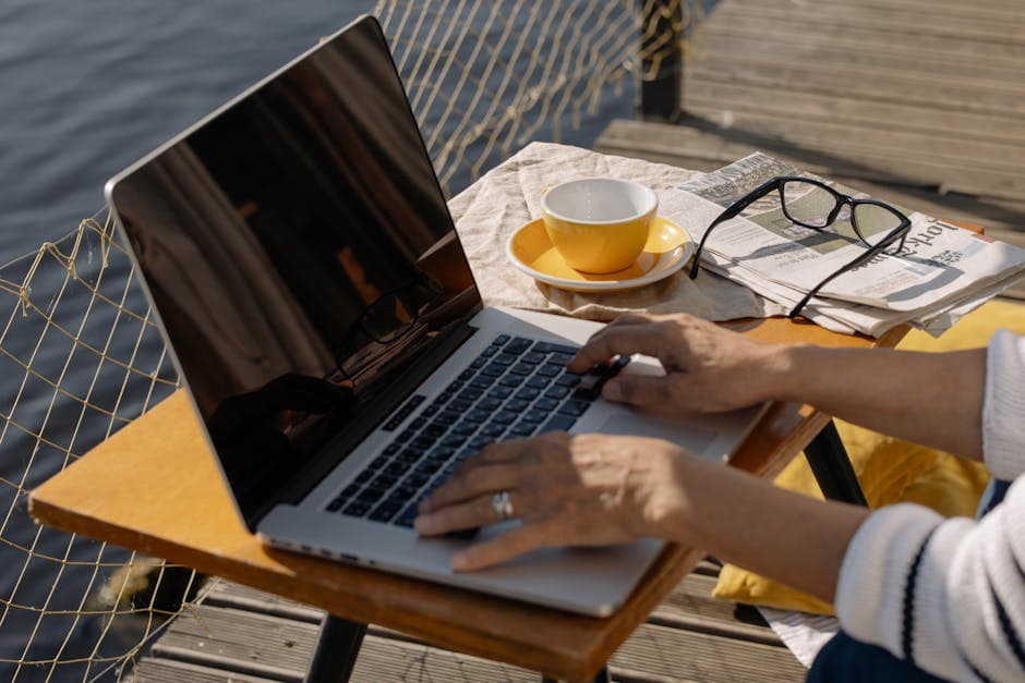 A person typing on a laptop outdoors by a lakeside with coffee and newspaper, ideal for remote work imagery.