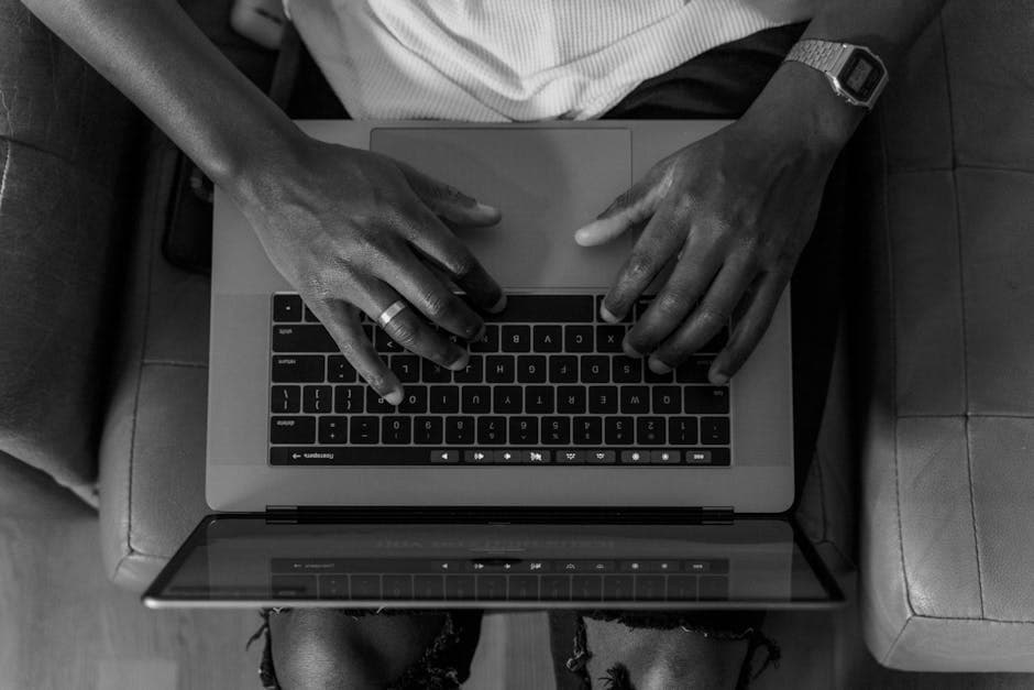 A black and white photo of hands typing on a laptop from an overhead perspective, conveying focus.