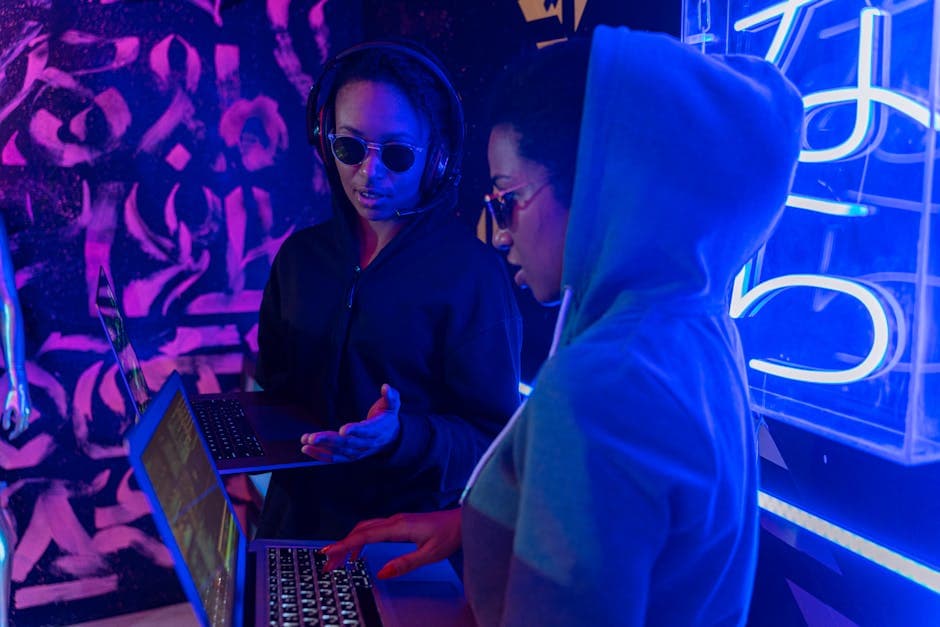 Two people using laptops in a neon-lit room, discussing technology concepts.