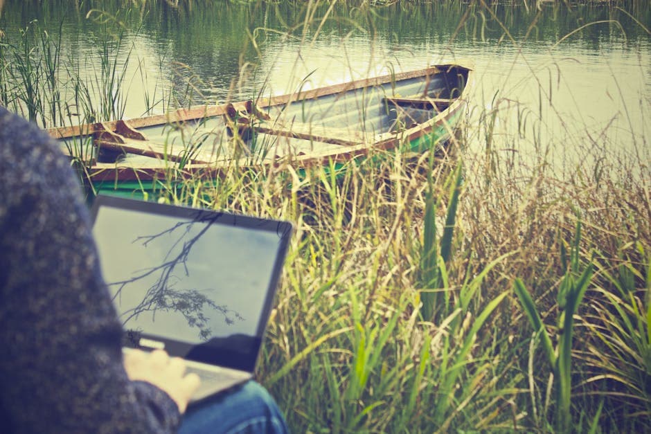 Person using laptop outdoors near a boat on a tranquil lake, showcasing remote work lifestyle.